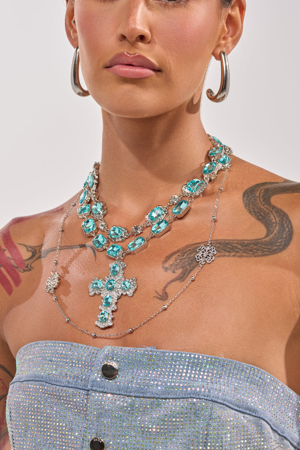A person wearing the REALLY WITH IT LAYERED NECKLACE—vintage-inspired turquoise gemstone strands with a turquoise cross pendant—pairs it with large hoop earrings, visible tattoos, and a strapless, sparkly light blue top. 1