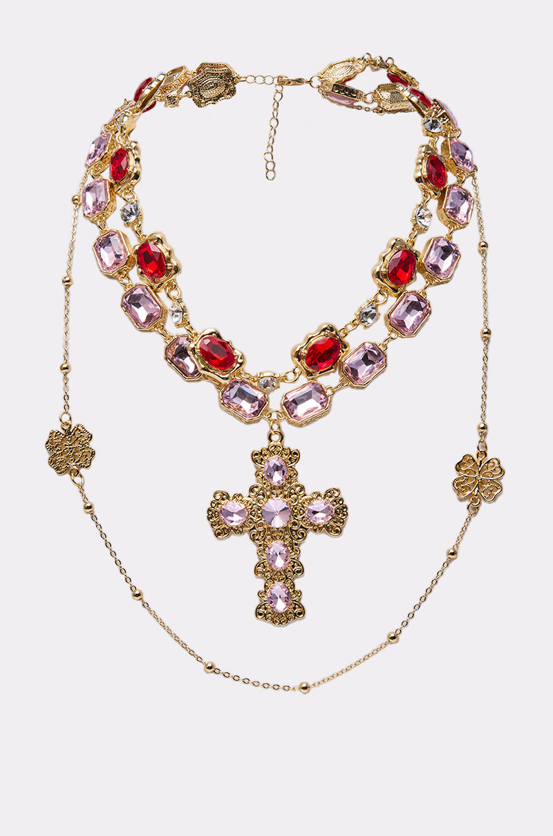 The LAYERED UP NECKLACE is a faux gold necklace featuring red and lavender gemstones, a striking cross pendant with lavender stones, floral gold accents, and layered chains adorned with ornate gold flower charms. 0
