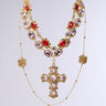The LAYERED UP NECKLACE features a faux gold chain with alternating large red and clear gemstones, highlighting a central bejeweled cross pendant. It's accented by two gold floral details on the dangling sections, set against a plain gradient background.