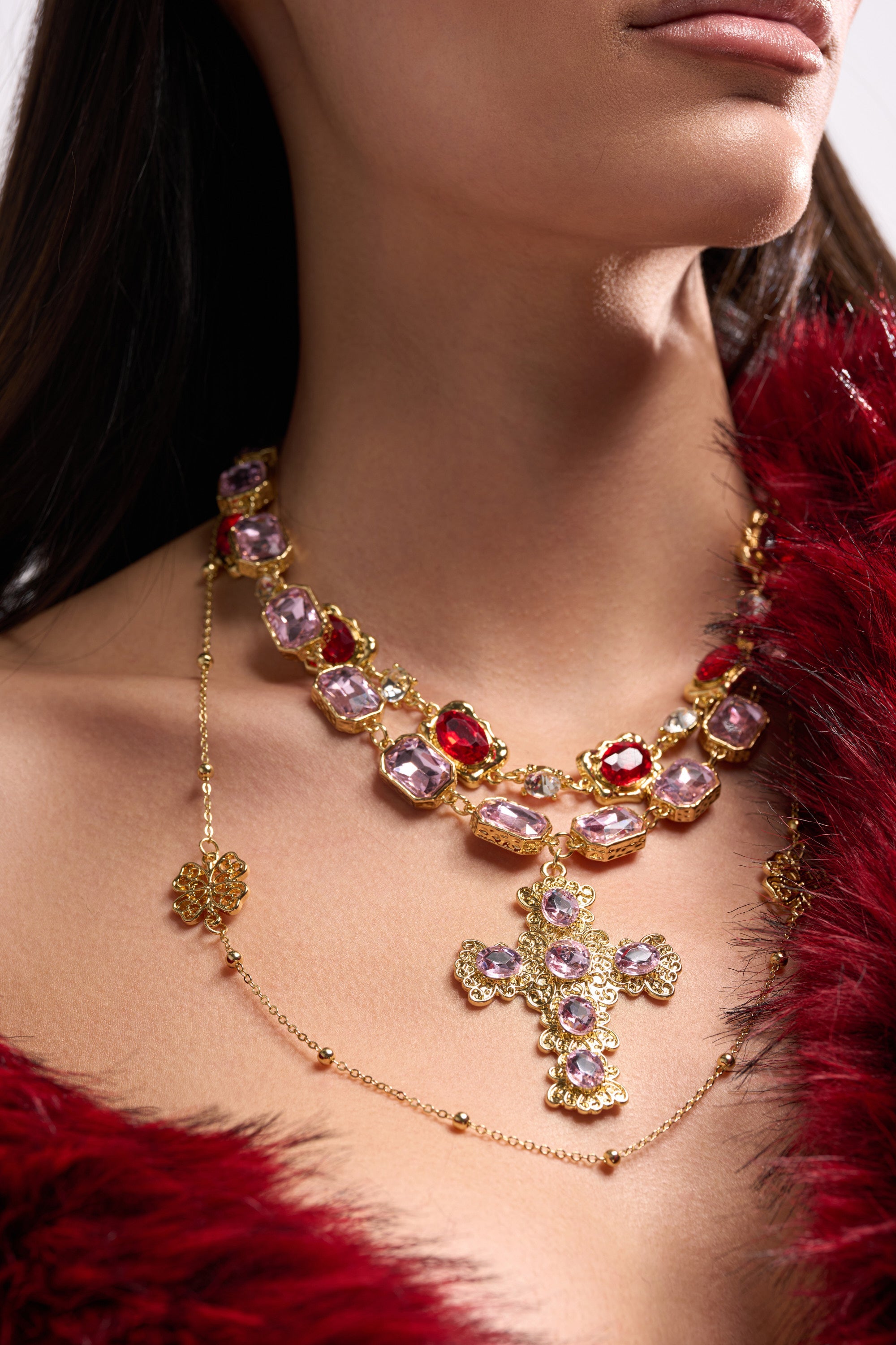 A woman wears the LAYERED UP NECKLACE featuring faux gold layers with purple and red stones, a cross pendant on a gem-encrusted gold chain, and a red fur garment draped over her shoulder. Her face is partially visible. 1