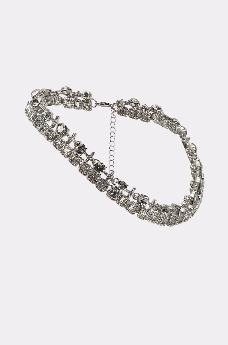 The BLING RING NECKLACE IN SILVER features a repeating pattern of sparkling, square-shaped crystals and a chain link extender, displayed against a plain white background. 2