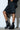 A person wearing knee-length black leather shorts with side laces pairs them with the AKI BLACK STRETCH PU CHUNKY BOOTIE and holds a small black handbag, all set against a white brick wall backdrop.