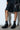 Dressed in a black leather ensemble, they stand before a white brick wall, showcasing their lower half. Their chic look is completed with a small black handbag and the AKI BLACK STRETCH PU CHUNKY BOOTIE.