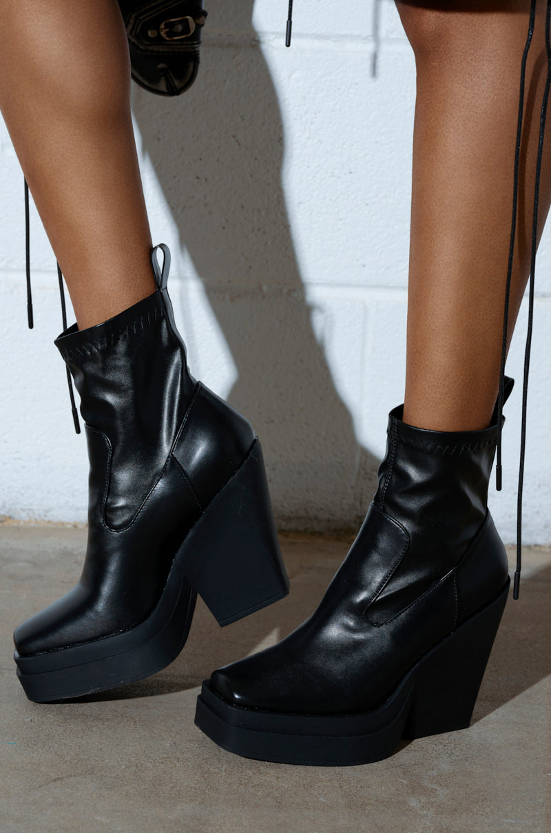 Someone stands on a concrete floor against a white wall, wearing AKI BLACK STRETCH PU CHUNKY BOOTIES. These vegan leather booties have thick platform soles and front zippers. The person's legs up to the knee cast shadows on the wall.