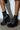 Someone stands on a concrete floor against a white wall, wearing AKI BLACK STRETCH PU CHUNKY BOOTIES. These vegan leather booties have thick platform soles and front zippers. The person's legs up to the knee cast shadows on the wall.