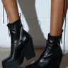 Someone stands on a concrete floor against a white wall, wearing AKI BLACK STRETCH PU CHUNKY BOOTIES. These vegan leather booties have thick platform soles and front zippers. The person's legs up to the knee cast shadows on the wall.