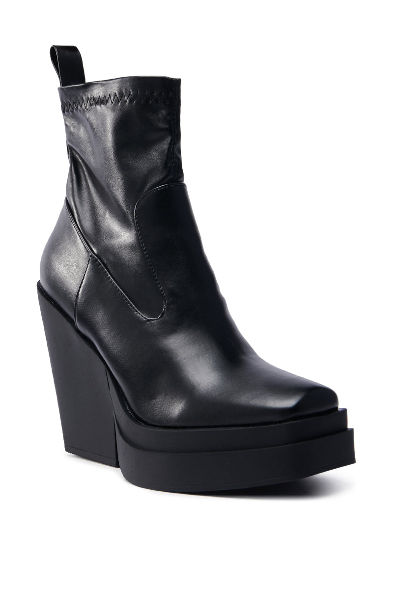 Introducing the AKI BLACK STRETCH PU CHUNKY BOOTIE, a vegan leather bootie with a thick high wedge platform sole, squared toe, visible stitching at the top, and a handy pull tab at the back, all set against a plain white background.