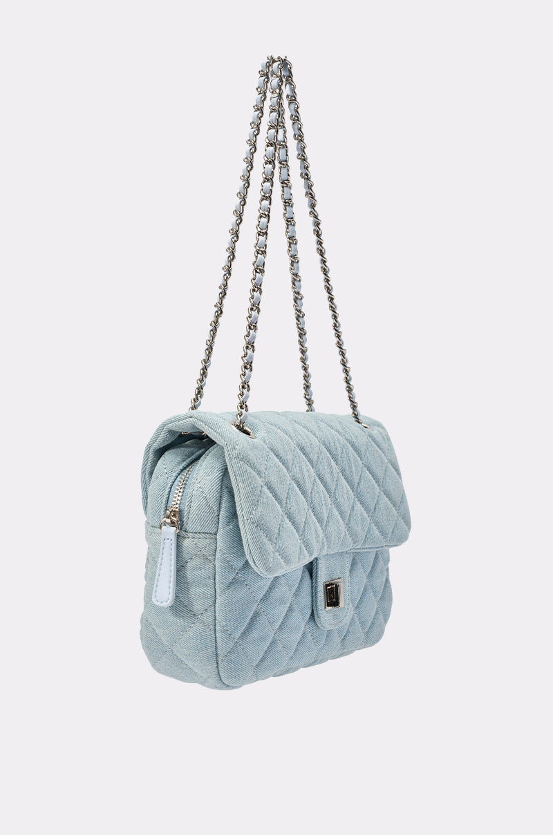 The BLUE JEAN BABY DENIM QUILTED PURSE features light blue quilted denim, a flap closure with silver turn-lock clasp, double chain straps, and a small white tag accenting the side for added style. 2