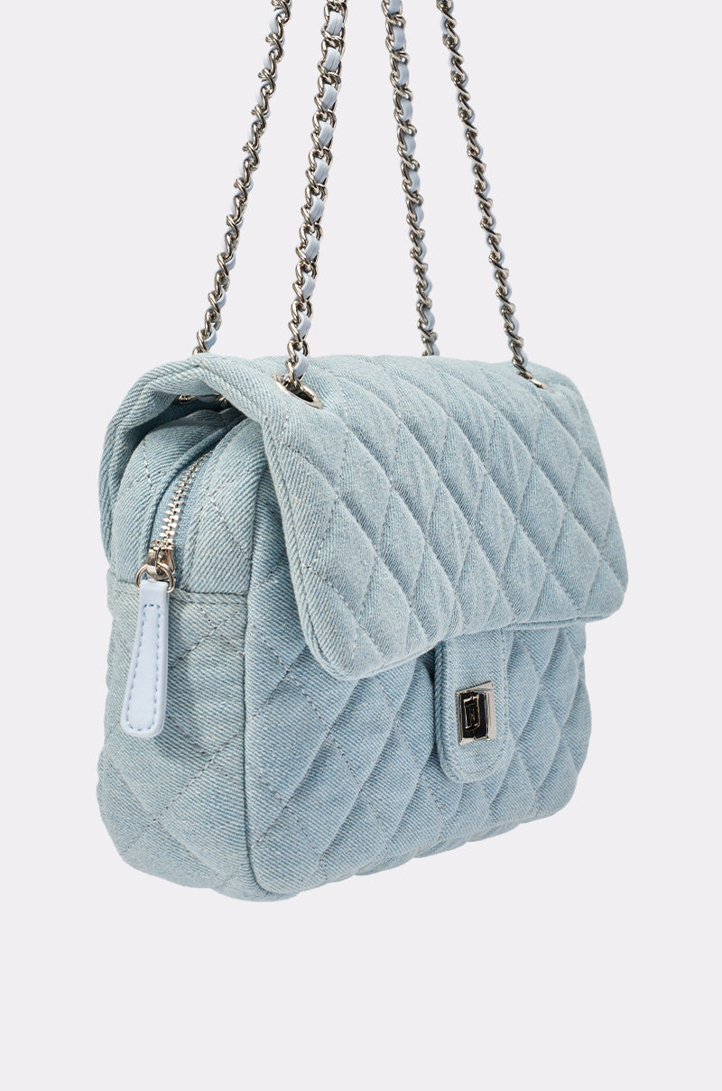 The BLUE JEAN BABY DENIM QUILTED PURSE is a light blue quilted denim bag with a flap, silver-tone turn-lock clasp, and silver chain strap woven with blue material. It features a side zipper and a structured rectangular shape. 3