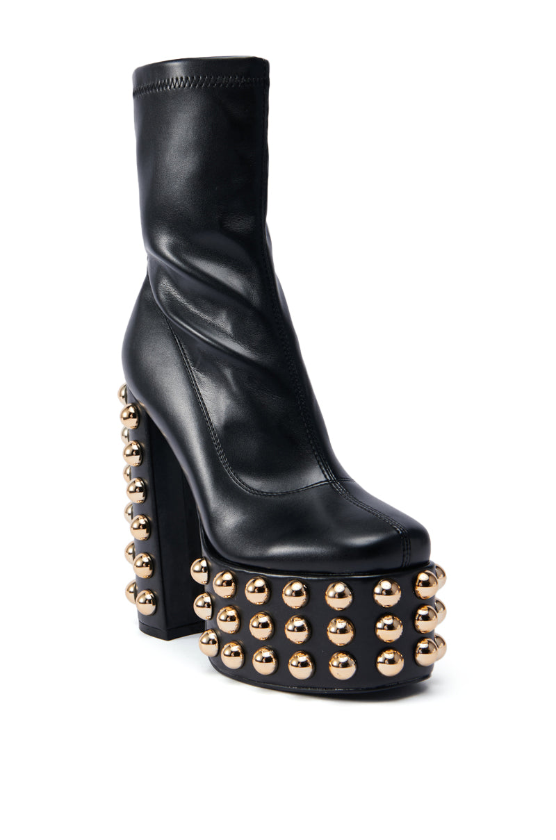 The AZALEA WANG AKITO CHUNKY BOOTIE is a trendy platform boot featuring a black high-heeled ankle design made from faux leather. It boasts a chunky platform and gold stud embellishments for a bold, decorative touch. The sleek upper section includes a rounded toe, while the studs adorn the heel and platform area.