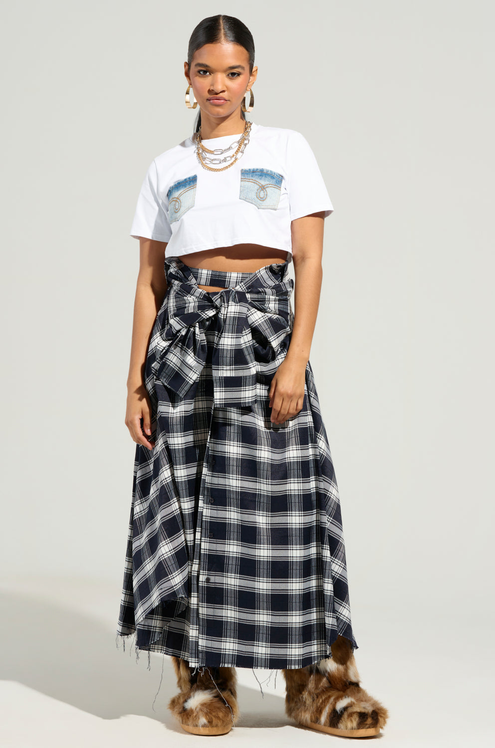A person stands against a plain background wearing a cropped white t-shirt featuring a graphic, the NEED YOU PLAID MAXI SKIRT adorned with a large bow and crafted from flannel, along with fur-covered boots. They have braided hair and wear layered necklaces.
