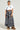 A person stands against a plain background wearing a cropped white t-shirt featuring a graphic, the NEED YOU PLAID MAXI SKIRT adorned with a large bow and crafted from flannel, along with fur-covered boots. They have braided hair and wear layered necklaces.