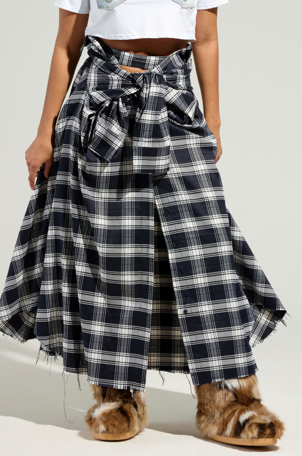 A person is wearing the NEED YOU PLAID MAXI SKIRT, featuring a large bow at the waist and a black, white, and gray checkered pattern crafted from flannel. They've paired it with a white top and ankle-high brown boots that boast fluffy textures.