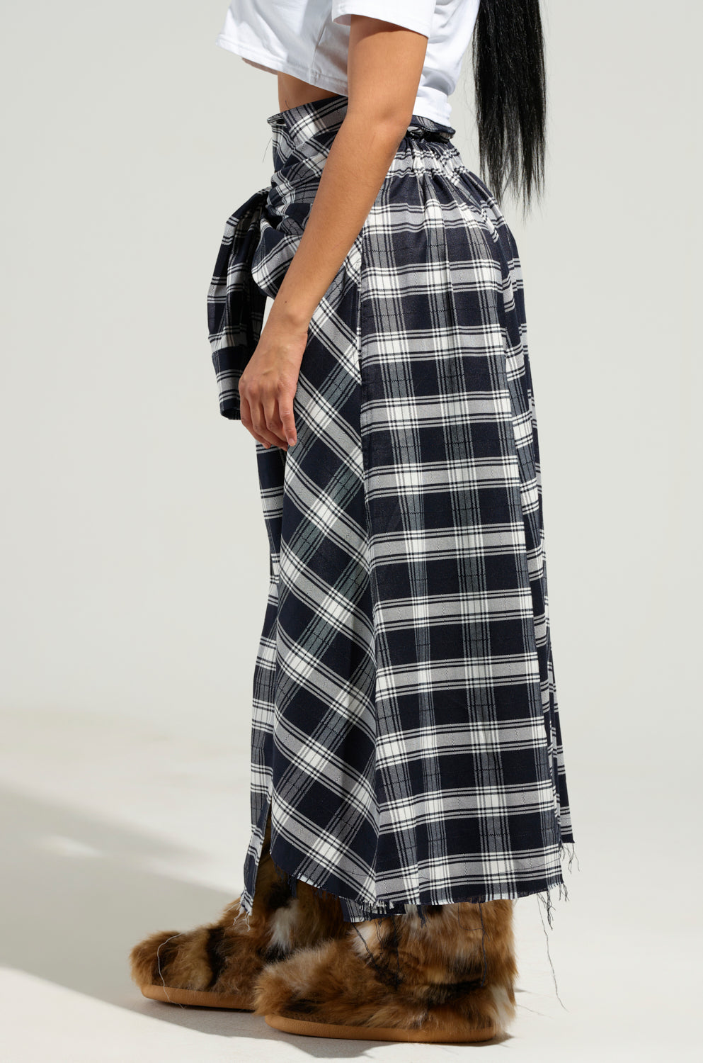 A person wearing a white top paired with the NEED YOU PLAID MAXI SKIRT, designed with flannel fabric and accented by a tied bow at the waist. Their furry brown slippers peek out as they stand sideways, long black hair cascading gracefully down their back.