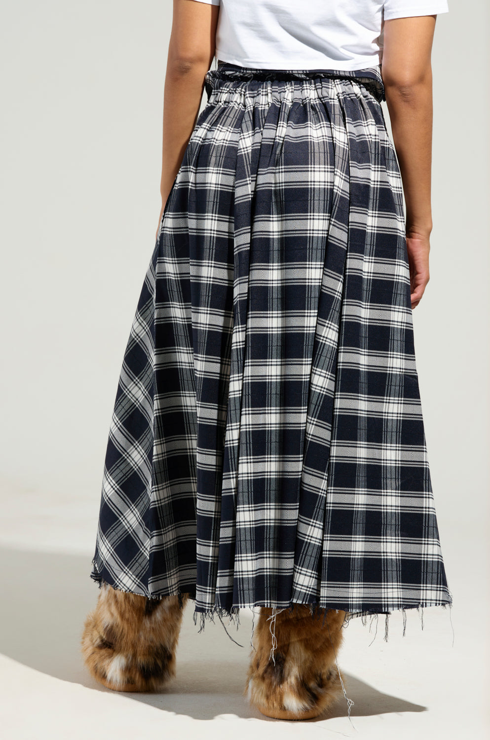 A person is seen wearing the NEED YOU PLAID MAXI SKIRT, a black and white plaid maxi skirt featuring a frayed hem and made from flannel. It is paired with a white shirt and brown furry boots in a side view against a plain background.