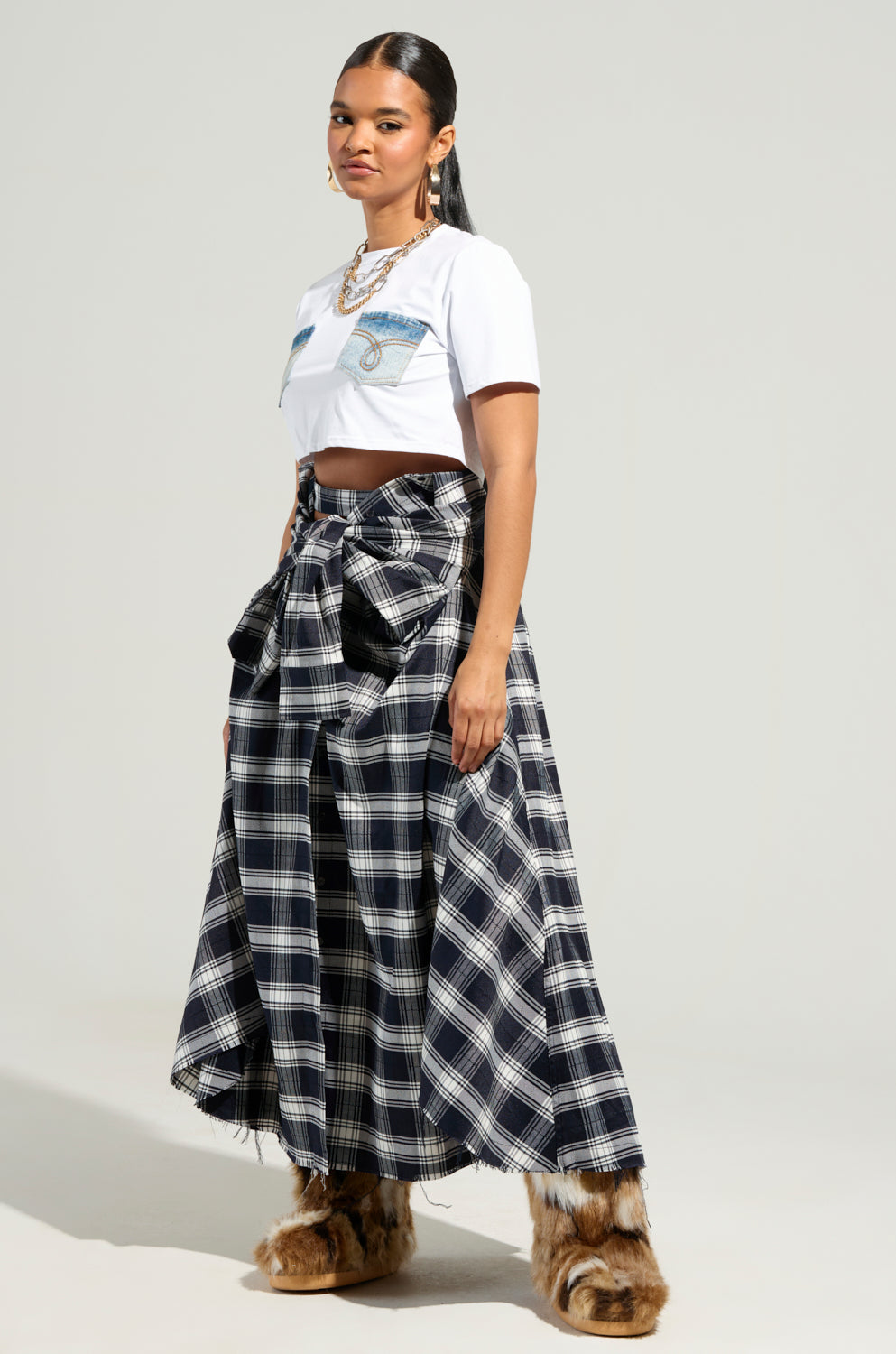 A person wears a white cropped t-shirt and the NEED YOU PLAID MAXI SKIRT featuring a high-rise silhouette adorned by a large bow, paired with furry brown boots. Long, dark hair cascades down their shoulders, accessorized with chunky gold necklaces. The backdrop is a plain light gray.