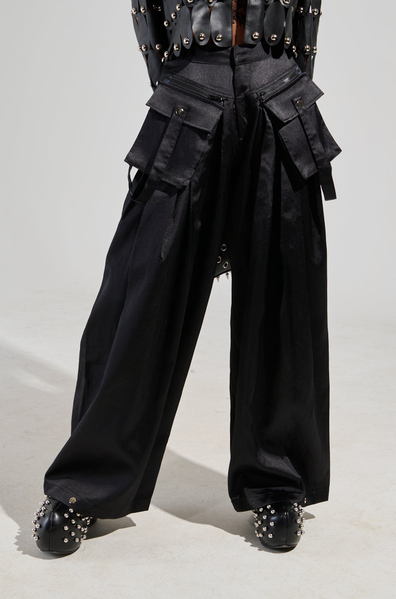 A person dressed in the A Moment Apart wide-leg trousers in black, featuring utility-style pockets and metal embellishments, paired with studded black shoes. The upper body is partially visible, showcasing a garment decorated with metal rivets against a plain and neutral background.