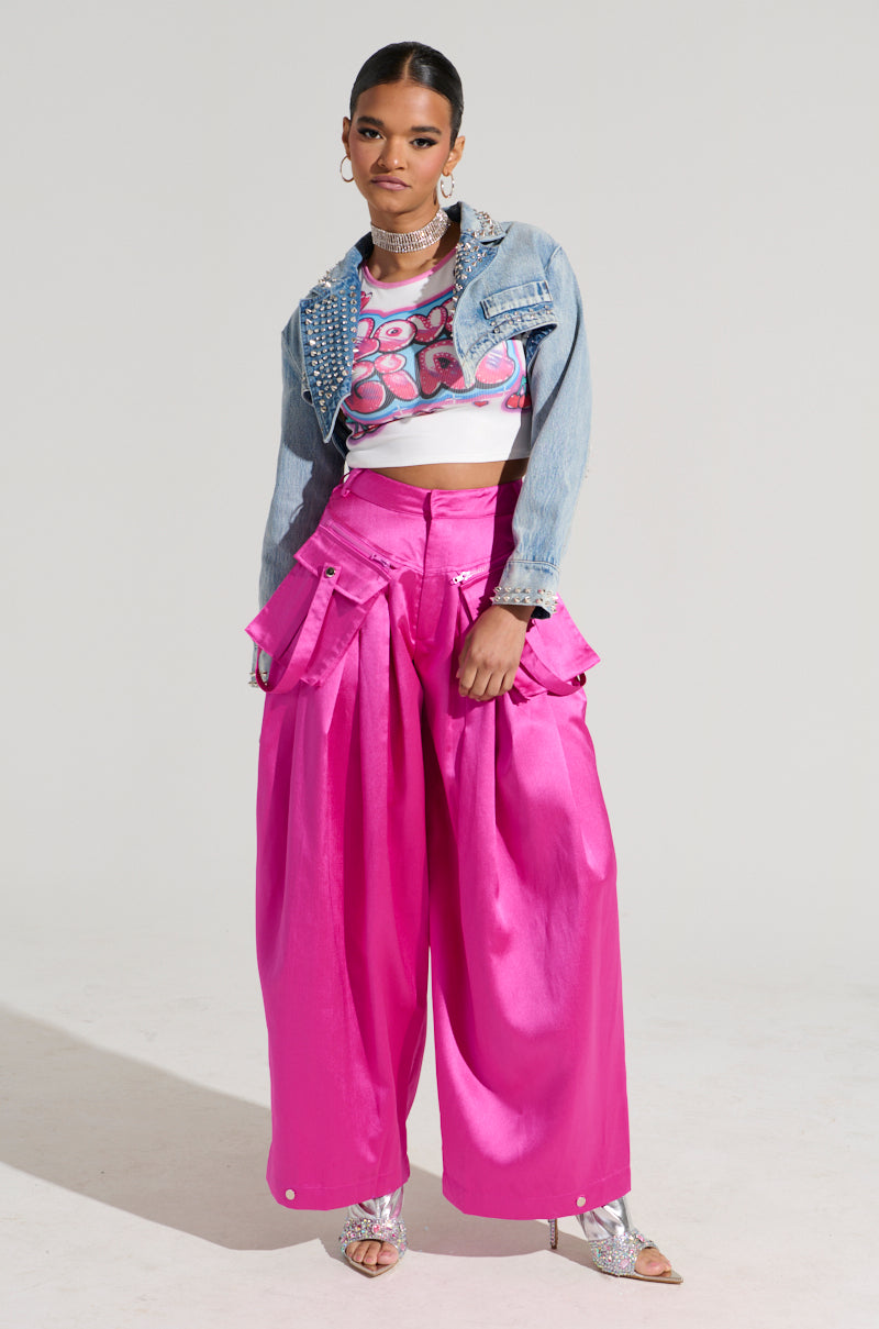 A model poses against a plain background wearing the vibrant pink A MOMENT APART WIDE LEG TROUSER WITH POCKETS from the AKIRA Label, paired with a cropped denim jacket adorned with studs and a colorful, patterned crop top underneath. She completes the look with silver sandals and bold jewelry.