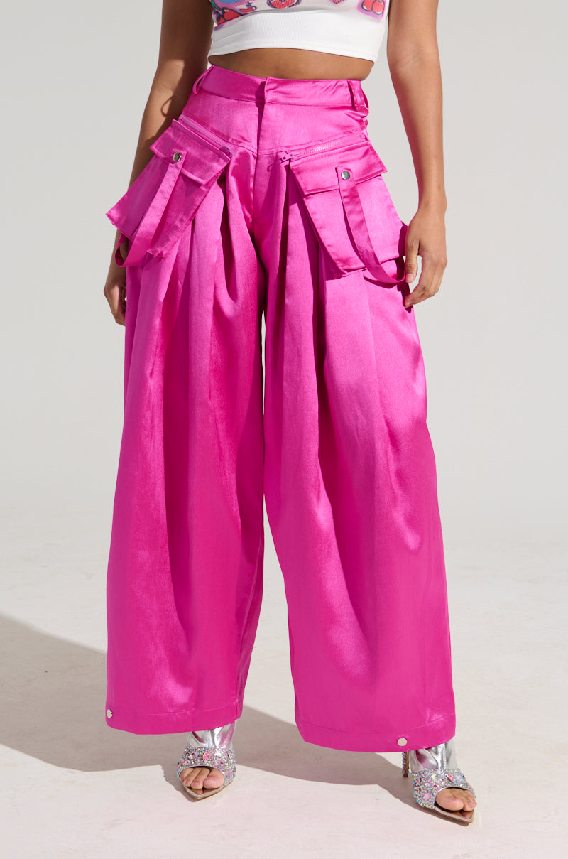 A person wearing the A MOMENT APART WIDE LEG TROUSER WITH POCKETS IN PINK from the AKIRA Label, featuring large cargo-style pockets and silver buttons, stands in a well-lit area. They also wear a crop top and silver, glittery high-heeled shoes. The outfit is bold and eye-catching against the plain white background.