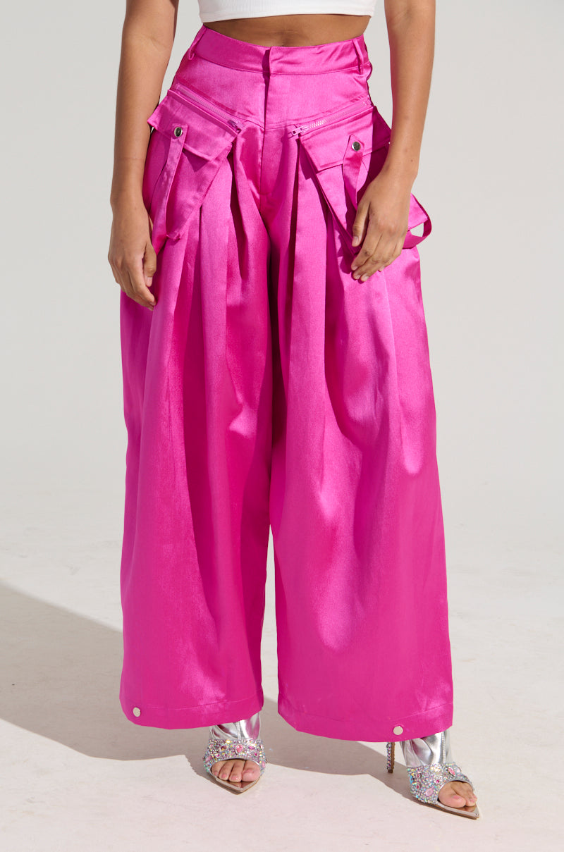 A person wearing the vibrant pink "A MOMENT APART WIDE LEG TROUSER WITH POCKETS IN PINK" by AKIRA Label, which features large pockets and a high waist. The trousers are paired with open-toe silver heels. The top part of the person's outfit is not fully visible.