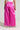 A person is wearing the vibrant pink "A Moment Apart Wide Leg Trouser with Pockets" from AKIRA Label, featuring a high-waisted fit and shiny fabric. The trousers have large pockets at the back, and the individual is paired with a sleeveless white top and glittering high-heeled shoes. They are standing with their back to the camera.