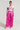 A woman stands against a plain background. She is wearing a white crop top with a colorful graphic design and the A MOMENT APART WIDE LEG TROUSER WITH POCKETS IN PINK from the AKIRA Label. She completes the look with strappy high-heeled sandals and a choker necklace.