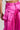 The person is wearing the A MOMENT APART WIDE LEG TROUSER WITH POCKETS IN PINK, which are vibrant silky pants with a shiny and smooth fabric that reflects light gracefully. These AKIRA Label trousers boast unique flap and zipper pocket designs, and the person's white top peeks out partially.