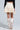 A person is shown from the back wearing a QUINN PLEATED TWEED MINI SKIRT in cream with pleats, paired with tall, shiny cream boots. They are also wearing a long-sleeve black top. The background is neutral and uncluttered.