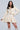 A woman with long dark hair is confidently posing in a cream-colored, textured two-piece outfit featuring the QUINN PLEATED TWEED MINI SKIRT and a matching cropped jacket. She completes the look with light-colored, knee-high boots.