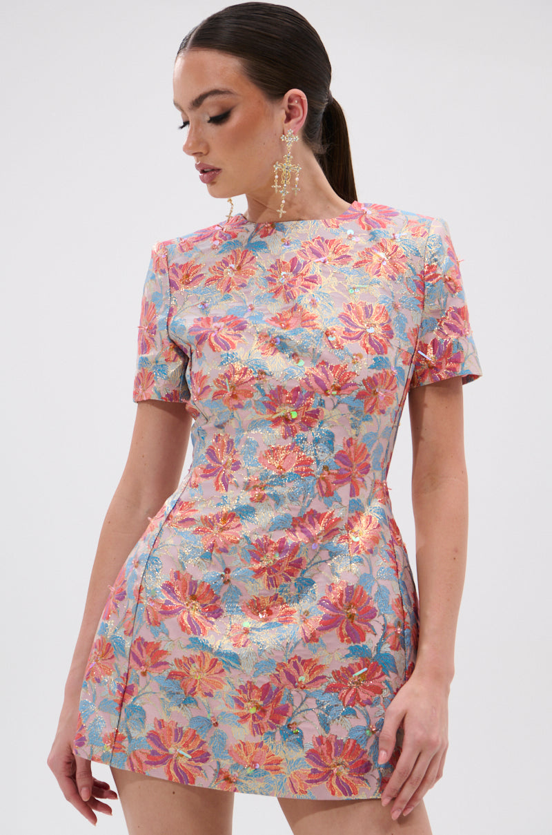 A woman in the PRETTY IN PINK BROCADE BEADED MINI DRESS with short sleeves and pastel floral brocade looks down to her left. Her long hair is in a ponytail, and she finishes the look with statement earrings.