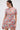 A woman in the PRETTY IN PINK BROCADE BEADED MINI DRESS with short sleeves and pastel floral brocade looks down to her left. Her long hair is in a ponytail, and she finishes the look with statement earrings.