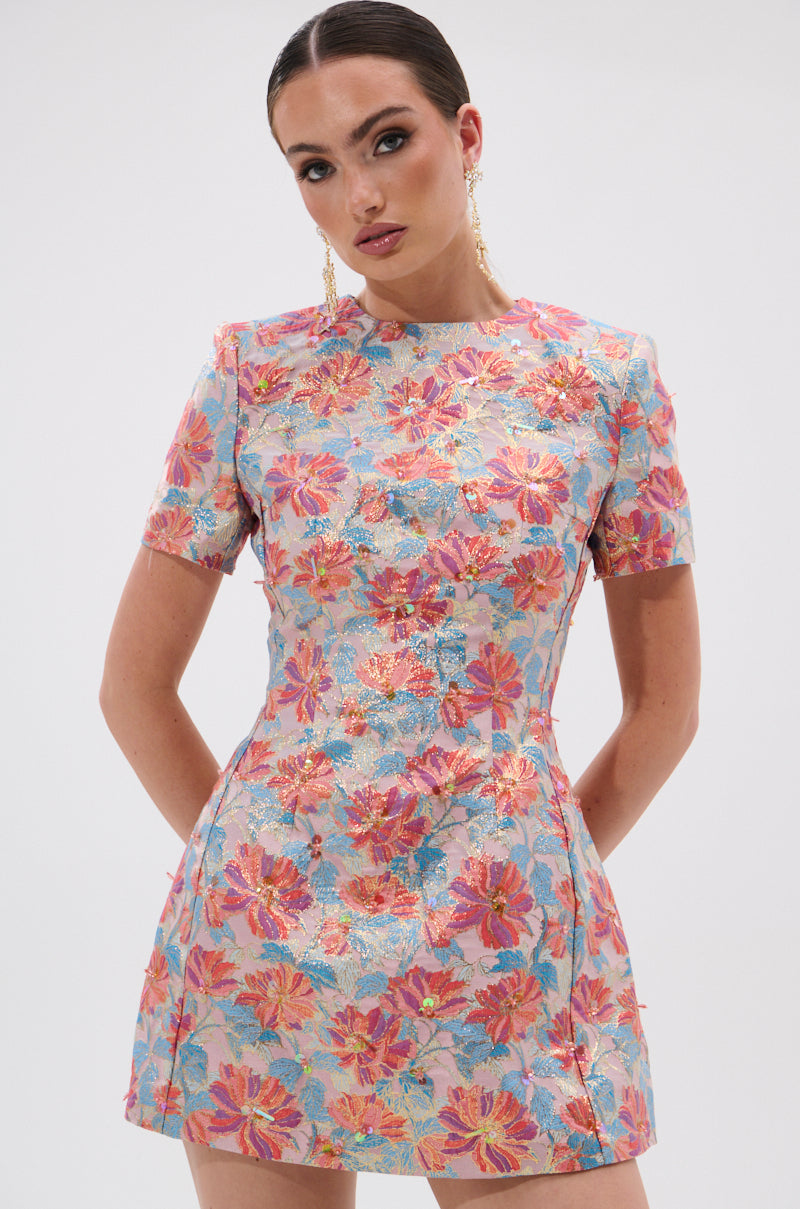 A woman showcases the PRETTY IN PINK BROCADE BEADED MINI DRESS, featuring short sleeves and a bold floral pattern. Her slicked-back hair, gold earrings, and striking makeup pop against a plain white background.