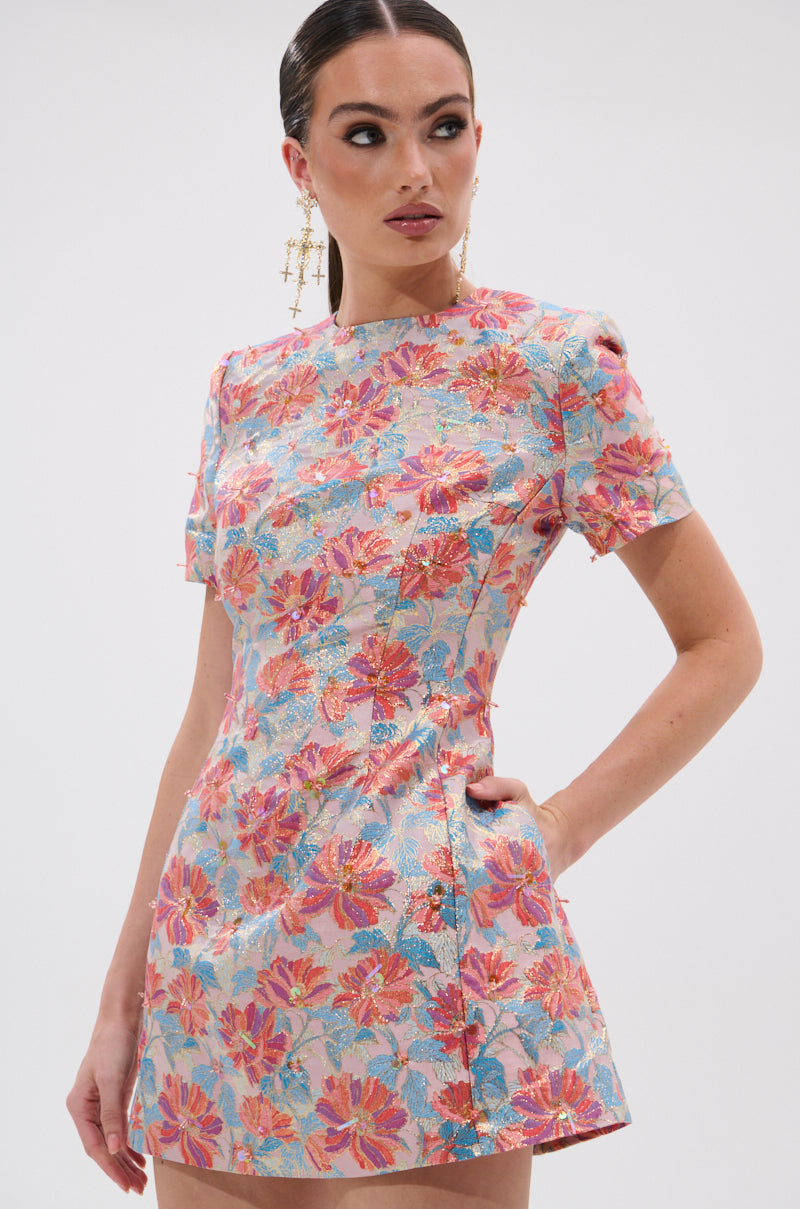 A woman models the PRETTY IN PINK BROCADE BEADED MINI DRESS, a fitted, short-sleeved mini dress with pink floral brocade. She wears statement earrings, has her hair pulled back, and stands with one hand in her pocket, gazing to the side.