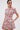 A woman models the PRETTY IN PINK BROCADE BEADED MINI DRESS, a fitted, short-sleeved mini dress with pink floral brocade. She wears statement earrings, has her hair pulled back, and stands with one hand in her pocket, gazing to the side.