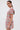 A woman with straight brown hair in a low ponytail wears the PRETTY IN PINK BROCADE BEADED MINI DRESS, featuring short sleeves and a colorful sparkling floral pattern. She stands sideways, facing the camera, accessorized with statement earrings.