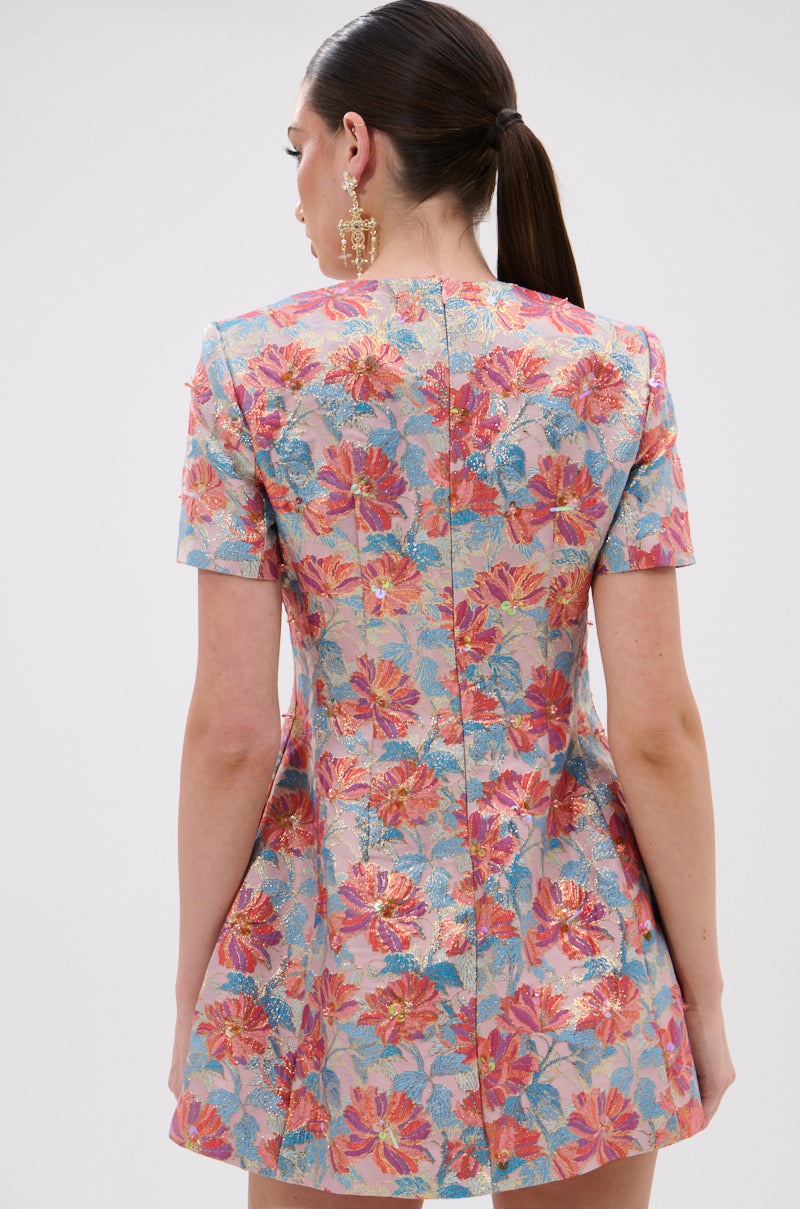 A woman with long dark hair in a ponytail models the PRETTY IN PINK BROCADE BEADED MINI DRESS, featuring short sleeves and a pink floral pattern. She faces away to highlight the back, accessorized with large gold earrings.