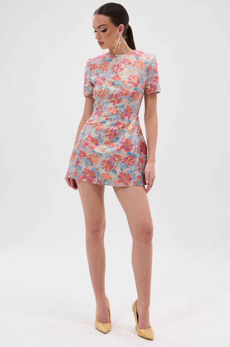 A woman models the PRETTY IN PINK BROCADE BEADED MINI DRESS—a short, fitted mini with colorful florals, short sleeves, and a high neckline—posing against a white background in gold heels and statement earrings.