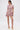 A woman models the PRETTY IN PINK BROCADE BEADED MINI DRESS—a short, fitted mini with colorful florals, short sleeves, and a high neckline—posing against a white background in gold heels and statement earrings.