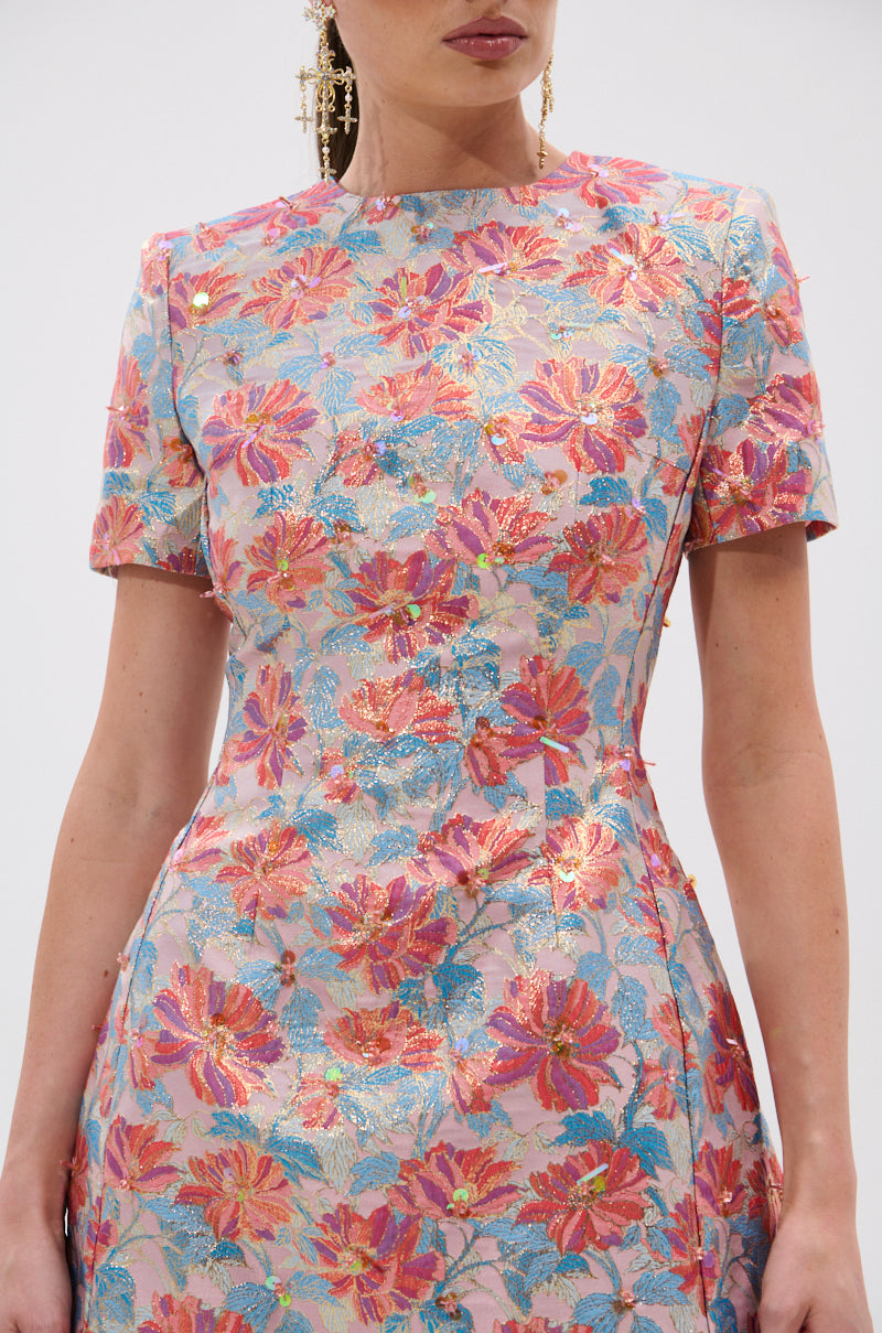 A woman wears the PRETTY IN PINK BROCADE BEADED MINI DRESS, featuring a vibrant pink, gold, blue, and red floral pattern with short sleeves and a high neckline. The image is cropped at her shoulders and knees.