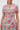 A woman wears the PRETTY IN PINK BROCADE BEADED MINI DRESS, featuring a vibrant pink, gold, blue, and red floral pattern with short sleeves and a high neckline. The image is cropped at her shoulders and knees.