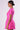 A woman stands in profile against a plain background, showcasing the Luxon Group Inc PRETTY IN PINK BROCADE BEADED MINI DRESS. The bright pink, textured dress features short sleeves, fits closely at the waist, and flares out slightly at the skirt. She has her hair pulled back in a neat bun and complements her look with pink drop earrings.