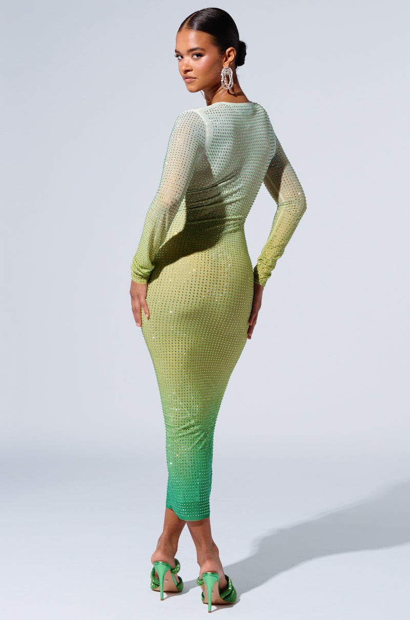 A woman stands posing, showcasing the back of her attire. She is adorned in the sophisticated Green With Envy Rhinestone Maxi Dress that features a long-sleeved, form-fitting silhouette crafted from a sheer double-lined mesh fabric that gradients from light to dark green. Her hair is pulled back, and she pairs the ensemble with large, dangling earrings and green high-heeled sandals.