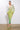 Against a plain white background, a model showcases the curve-hugging GREEN WITH ENVY RHINESTONE MAXI DRESS. This form-fitting, long-sleeved dress features an enchanting green ombré colorway adorned with sparkling rhinestones and a deep neckline with a delicate tie. She completes her look with green embellished heeled sandals.