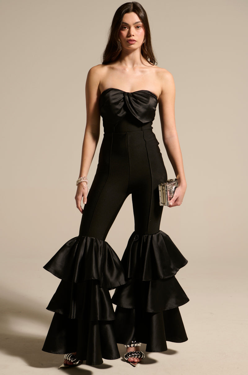 A woman wearing the "I JUST WANNA DANCE FLARE LEG JUMPSUIT," featuring tiered ruffle pants and a sweetheart neckline, stands against a plain background. She holds a small clutch and wears bracelets, with her loose hair cascading past her shoulders, embodying elegance.