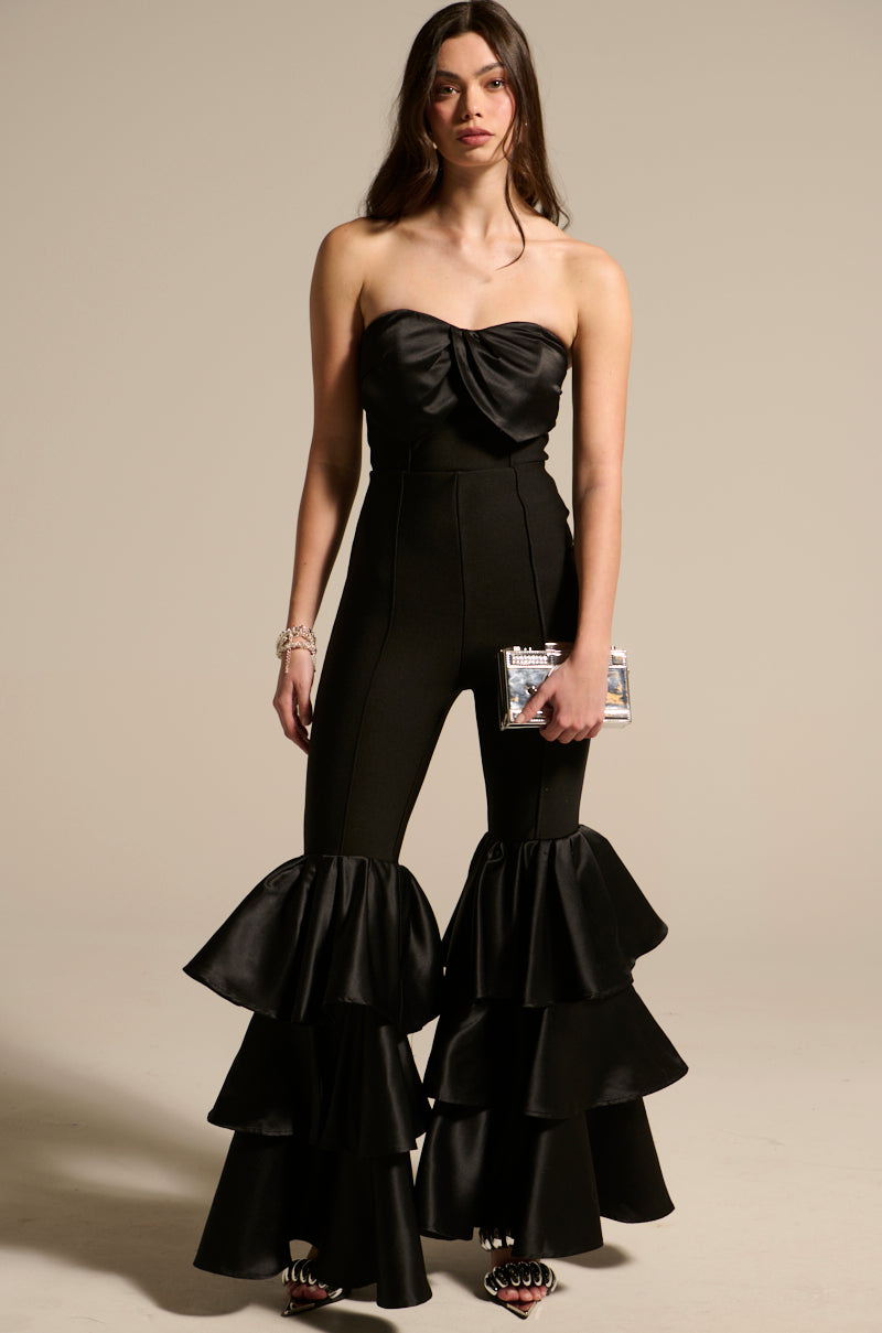 Wearing the "I Just Wanna Dance Flare Leg Jumpsuit," someone exudes confidence in a strapless black silhouette with ruffled flares, paired with a small purse and high-heeled shoes offering super stretchy comfort, all set against a plain backdrop.
