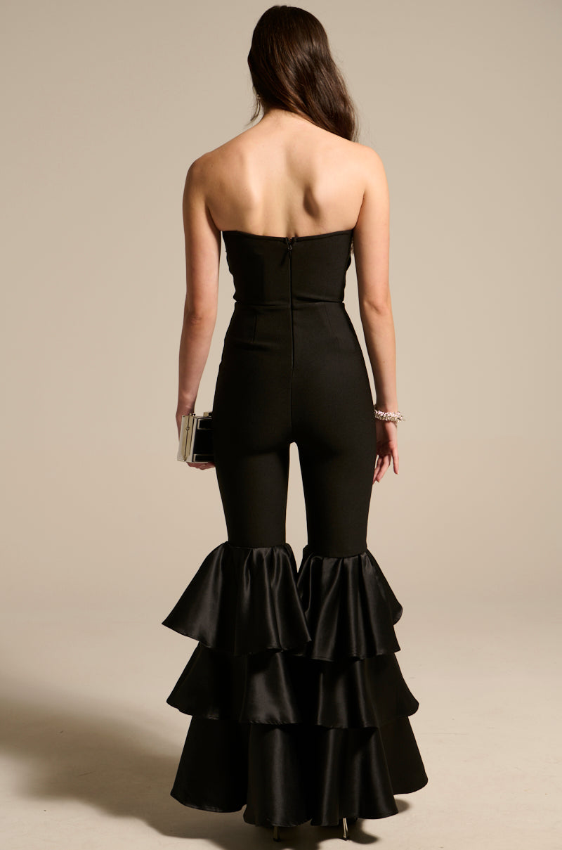Rear view of a woman in the I JUST WANNA DANCE FLARE LEG JUMPSUIT, strapless and black with flare leg bottoms. She holds a clutch, her long brown hair cascading down her back against a plain, neutral background—ideal for the dance floor.
