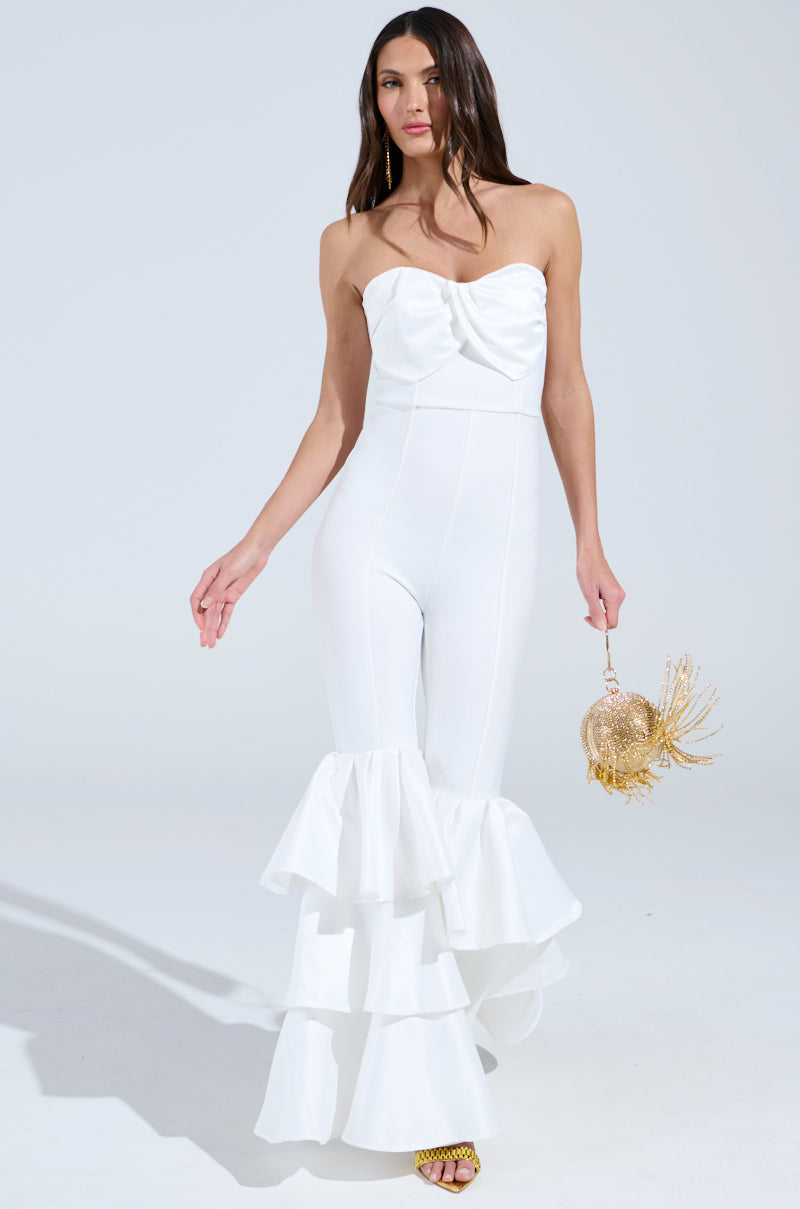 A woman exudes confidence in the I Just Wanna Dance Flare Leg Jumpsuit in White, showcasing a strapless neckline and layered ruffle bottoms. She accessorizes with a small gold decorative purse and yellow heels against a light gray backdrop.