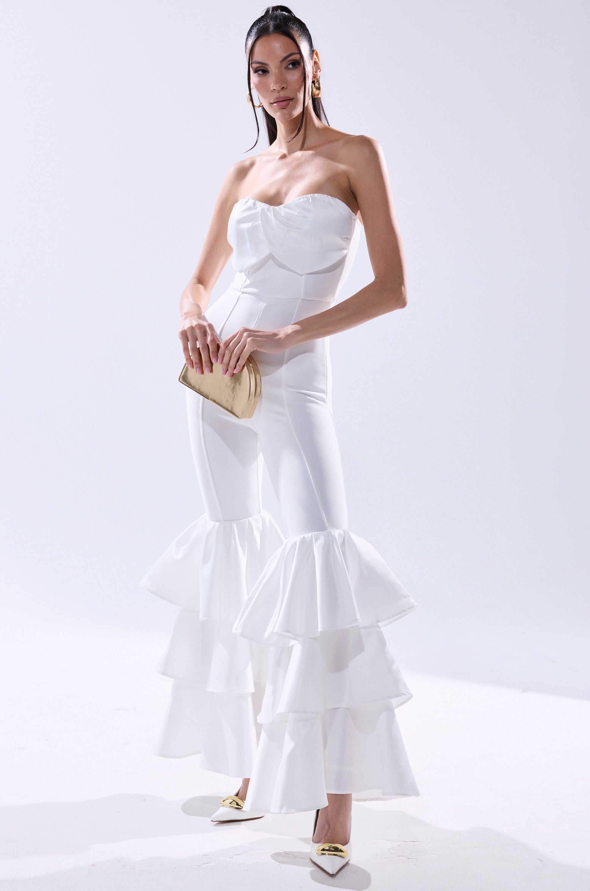 A woman stands against a light background wearing the I JUST WANNA DANCE FLARE LEG JUMPSUIT IN WHITE. She holds a beige clutch, wears pointed-toe heels, and her straight hair is tucked behind her ears. 1