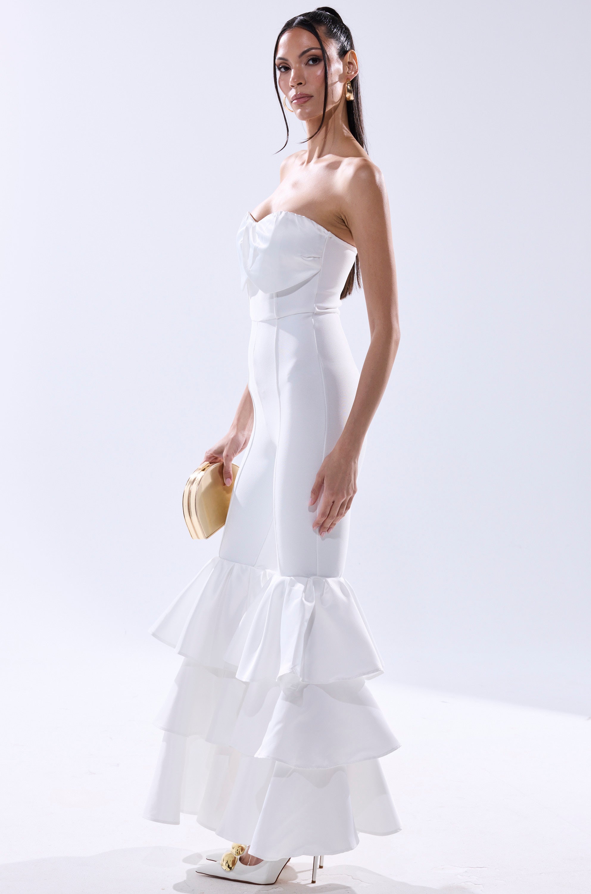 A woman stands in profile wearing the I JUST WANNA DANCE FLARE LEG JUMPSUIT IN WHITE, featuring a strapless neckline and tiered hem, styled with white heels and a petite beige clutch against a plain white background. 2