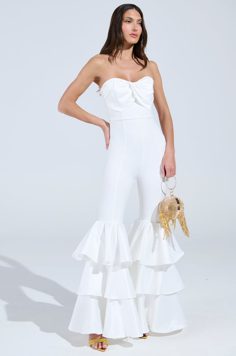 A woman stuns in the "I Just Wanna Dance Flare Leg Jumpsuit in White," featuring layered ruffles and a large bow at the bust. She accessorizes with a gold sequin fish-shaped handbag and strappy gold heels against a plain, light gray background.